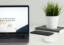 Photo of a laptop screen with the words WORKHARD ANYWHERE, a phone, mouse, plant, and a notebook
