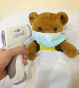 Photo of a teddy bear tucked in bed, wearing a mask, and with a hand holding a temperature sensor pointing at the bear.
