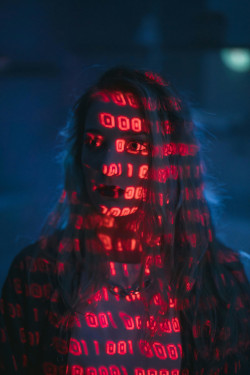 Altered photo of a white woman with long hair, dark blue background, with digital 001's in red superimposed over her.