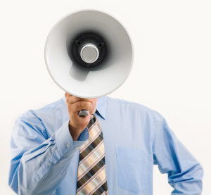 Photo of a man in a blue shirt and striped tie, holding a megaphone in front of his face.