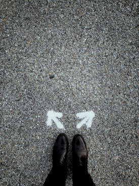 Grayscale photo of the lower portion of a man's legs, in black slacks and highly-polished black shoes, on a gray pebbled walkway, with two white chalk arrows in front of the toes,pointing in different directions.