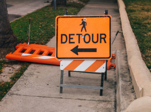 Photo of an orange Detour sign in the middle of a sidewalk.