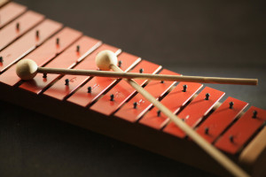 Photo of a segement of a dark red xylophone with two xylophone hammers crossed on top, on a dark-gray background.