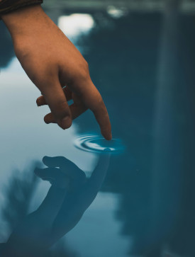 Photo of a hand reaching down with one finger to touch water; the hand is reflected, and a small ripple spreads from the finger-touch.