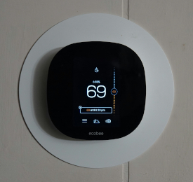 Photo of a black, rounded-square thermostat reading "69" on a white mounting circle and a cream-colored wall