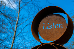Photo of a rusty sculpture of a hoop with a circular insert with "Listen" cut out, against a blue sky with a leafless tree to the left.