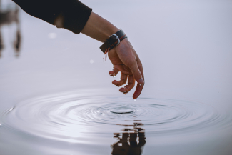 Photo of an arm reaching from top left toward water, with a ripple spreading from the forefinger and the hand reflected in the water. The arm wears a black sleeve and a black-banded wristwatch.